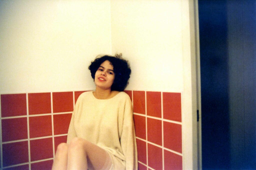 Sur la photographie couleur une femme aux cheveux courts et noirs est adossée aux deux murs d'une salle de bain aux carreaux orange. Elle porte un long pull et des bas blancs.