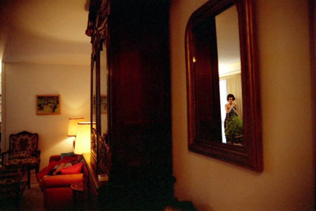 Sur la droite de la photographie couleur on voit de très loin le reflet d'une femme dans le miroir accroché au mur. Elle est presque invisible car la pièce est sombre et que le miroir est de profil. Dans la partie gauche de l'image on devine, à peine éclairée par une lampe, l'intérieur d'un salon bourgeois. Fait partie de la série des autoportraits photographiques argentiques.