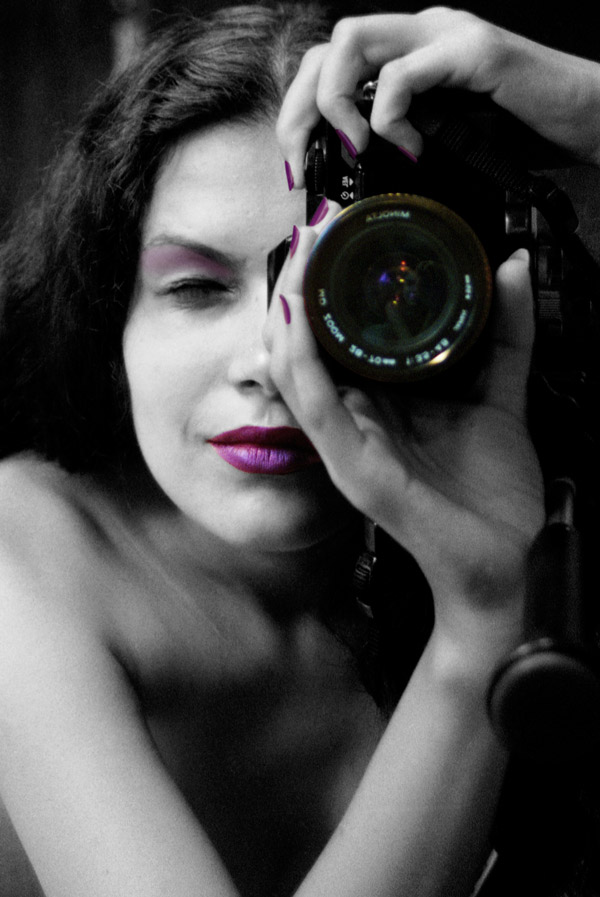 Portrait d'une femme brune, les yeux fermés, tenant verticalement un appareil photographique sur son œil gauche. Cette photo a été retouchée afin de paraître noire et blanche. Seuls sont restés en couleurs : le reflet dans l’objectif, les lèvres, les paupières et les ongles de la femme. Tous sont violet - rose.