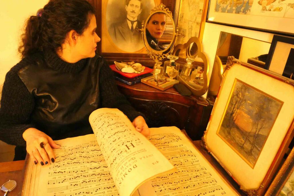 Femme brune en pull noir, laine et cuir, assise à un bureau. Elle est entourée de tableaux, de photos et de bibelots. Elle est face à nous et feuillette un antiphonaire. Mais elle tourne son visage vers le miroir qui est placé derrière elle. On voit donc son visage de profil et de face dans le reflet du miroir. La photo noire et blanche placée derrière le miroir est celle d’un homme moustachu en costume cravate. C’est une grande et très vieille photo. Le reflet de la femme est à côté de celui de l’homme du portrait.