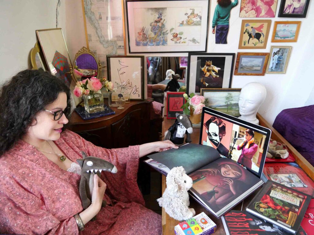 Femme de profil assise à un bureau. Avec trois peluches, elle regarde un écran d’ordinateur sur lequel apparaît un dessin représentant Raphaël Colantonio et une photographie d’Ava Gore. La femme brune, en robe d’intérieur rose orangé, est entourée de livres et de cadres contenant dessins ou photographies.