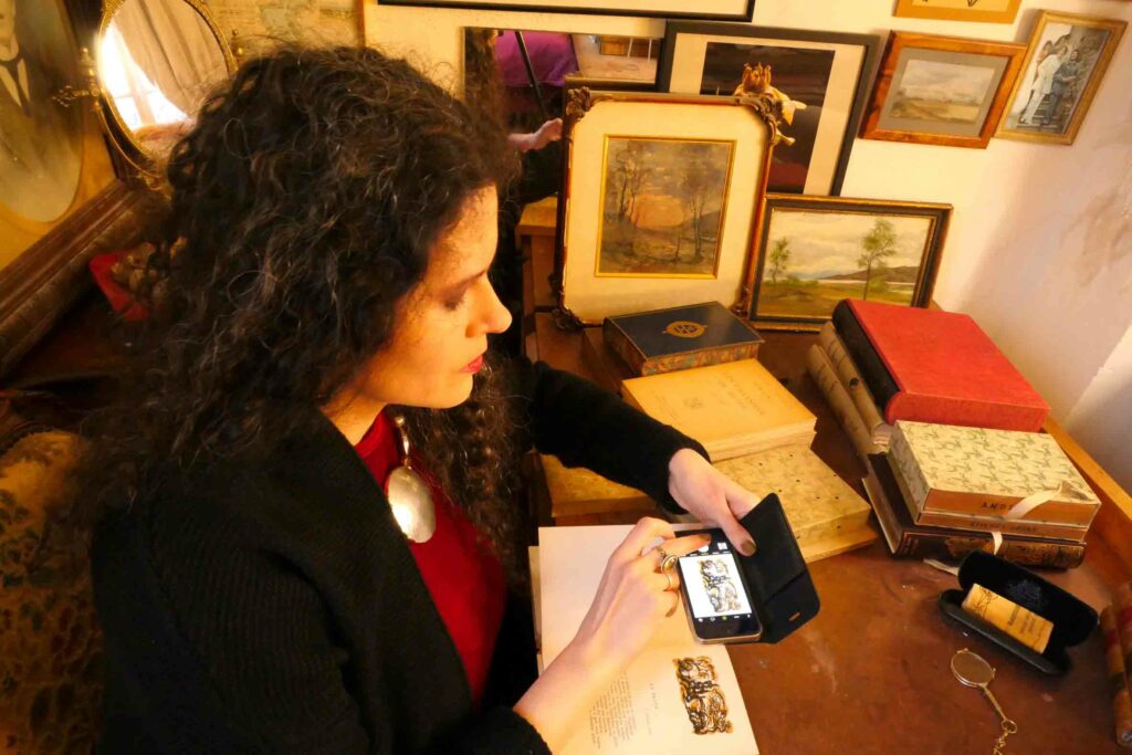 Femme brune en robe rouge et gilet noir assise, de profil, à un bureau. Elle est entourée de livres et de cadres contenant dessins ou photographies. Avec son téléphone portable elle photographie une illustration dans un livre.