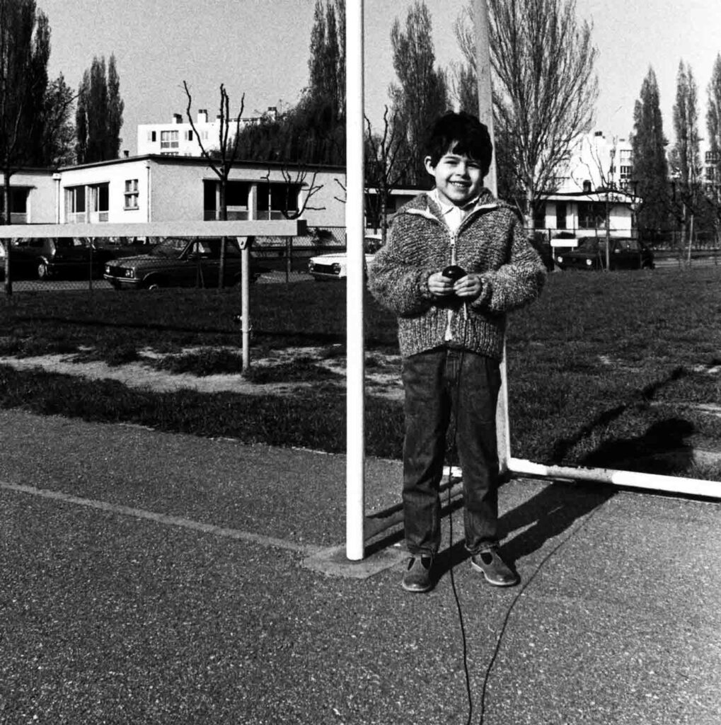 Photographie en noir et blanc prise à l'extérieur. Une petite fille brune avec des cheveux courts, pantalons et gilets, se tient debout dans les buts d'un terrain de foot. Elle sourit à l'objectif et tient dans ses mains la poire de l'appareil photographique.