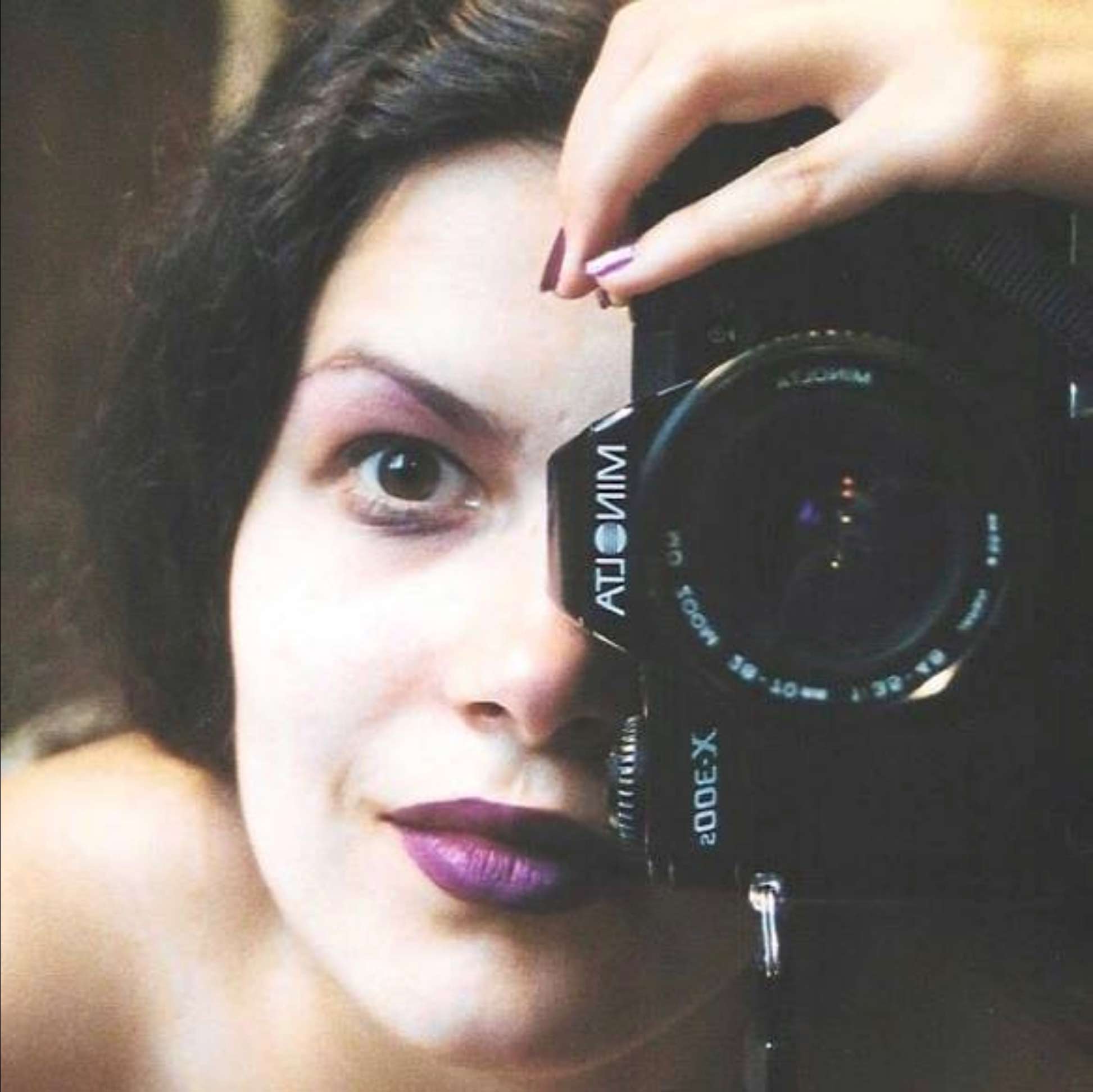 Portrait photographique en couleur. Gros plan sur le visage d'une jeune femme aux yeux et cheveux bruns. Son maquillage est violet. Elle tient verticalement son appareil photographique. L'objectif est devant et à la hauteur de son œil gauche.