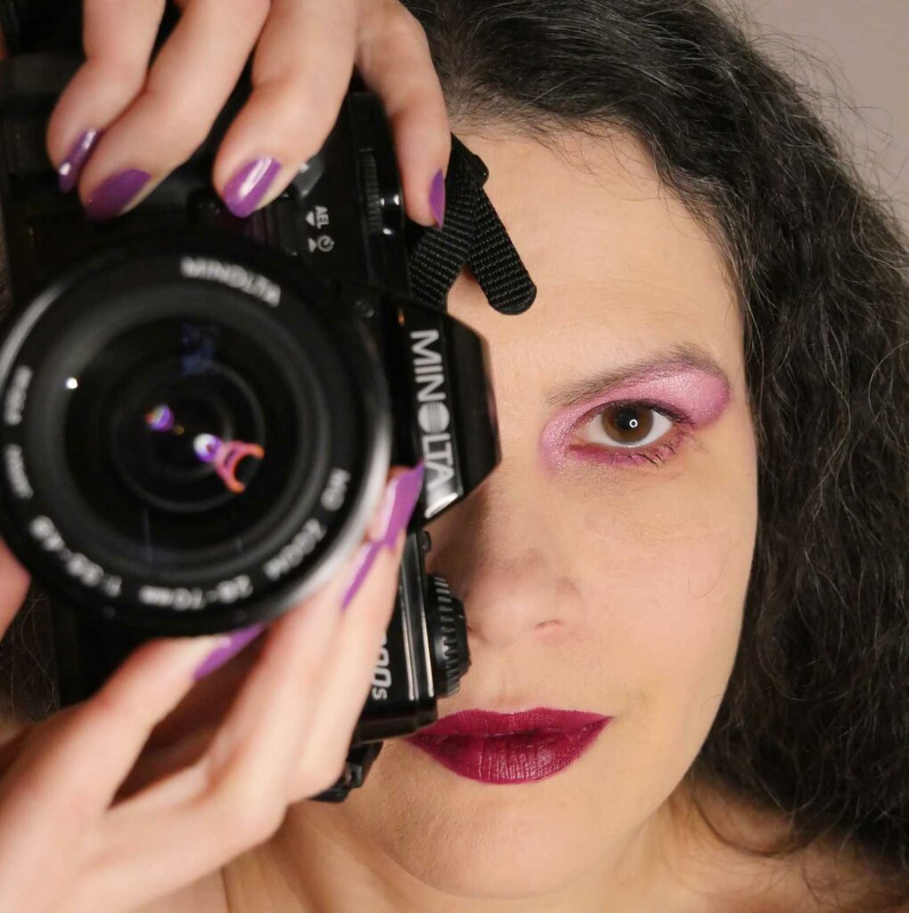 Portrait photographique en couleur. Gros plan sur le visage d’une femme aux yeux et cheveux bruns. Son maquillage est pourpre. Elle tient verticalement son appareil photographique Panasonic. L'objectif est devant et à la hauteur de son œil droit.