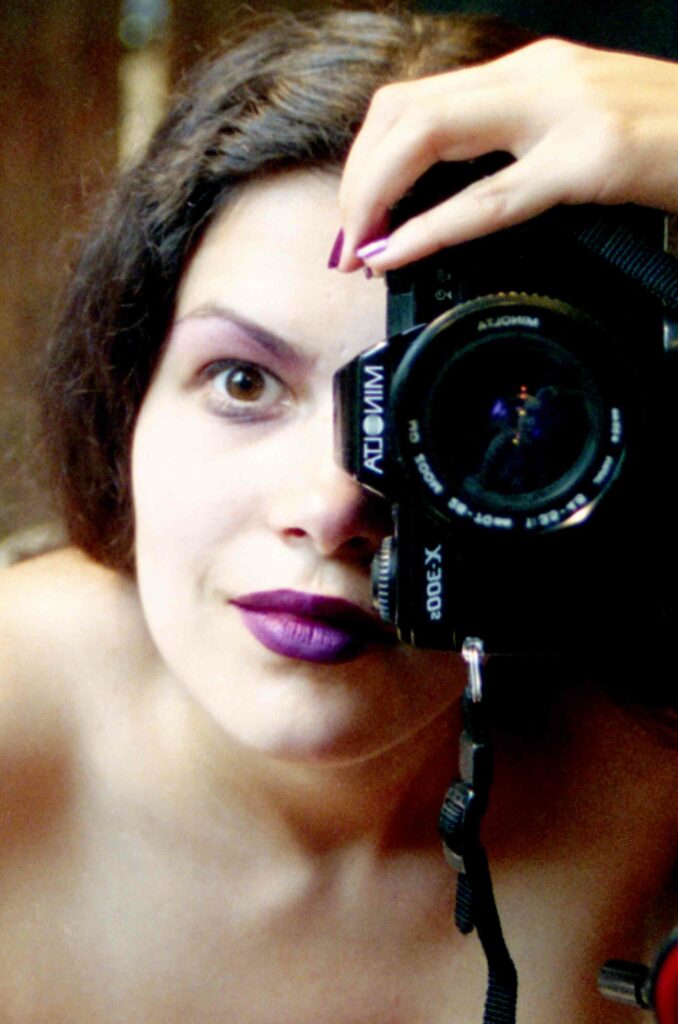 Portrait photographique en couleur. Gros plan sur le visage d'une jeune femme aux yeux et cheveux bruns. Son maquillage est violet. Elle tient verticalement son appareil photographique. L'objectif est devant et à la hauteur de son œil gauche.