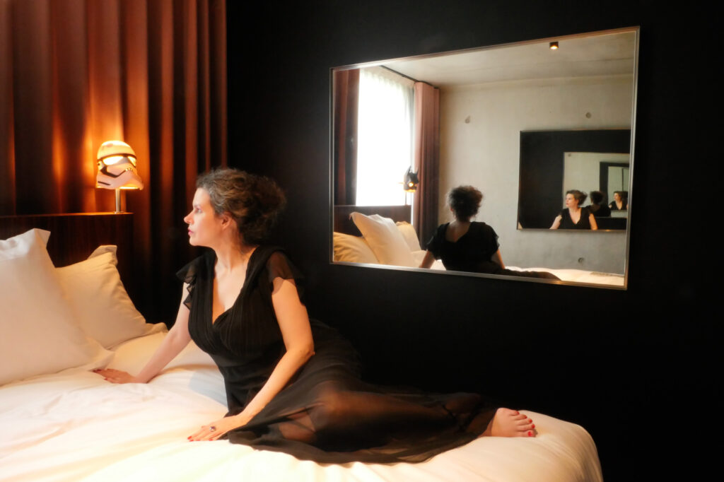 Photographie aux tons noir et orange dans une chambre d’hôtel. Une Femme en robe de soirée noire est allongée sur un lit blanc le long d’un miroir. Celui-ci reflète son dos et un second miroir. Les deux placés en vis-à-vis créent une mise en abyme dans l’image.